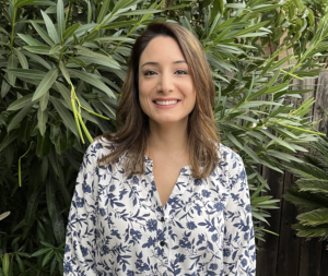 Woman smiling, wearing a blue and white floral blouse, standing in front of greenery.