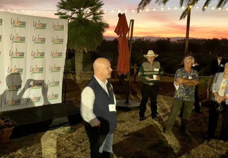 Man speaking at an outdoor event with a backdrop, other attendees, and sunset.