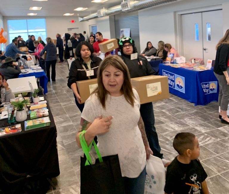 People carrying boxes at an event with tables displaying materials; a young child looks on.