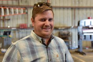 Man in plaid shirt smiling in a workshop, sunglasses on head.