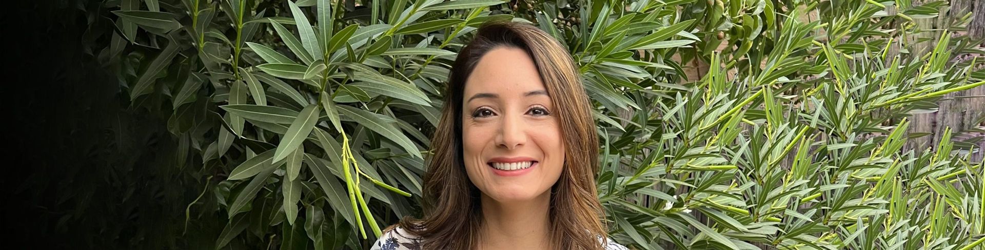 A woman smiles in front of green foliage. She has long brown hair and is wearing a patterned top.