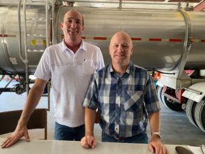 Two men stand near a large metal tank truck. One wears a white polo, the other a plaid shirt.