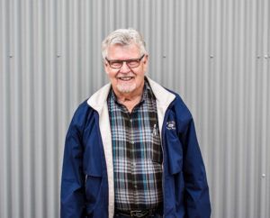 Man in glasses, blue jacket, and plaid shirt smiles in front of a corrugated metal wall.