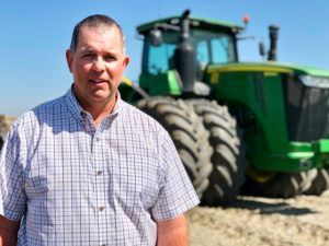 Man in button-down shirt stands near a large green tractor in a field under a blue sky.