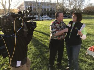 A man interviews a woman in a grassy yard, filmed by a camera. A large house is in the background.