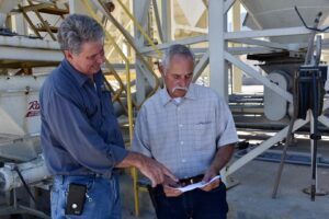 Two men reviewing paperwork near industrial equipment. One points.