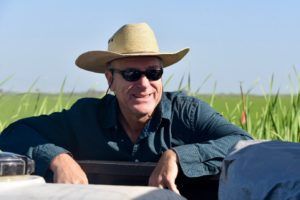Man wearing a straw hat and sunglasses leaning on something outdoors, smiling in front of a field.
