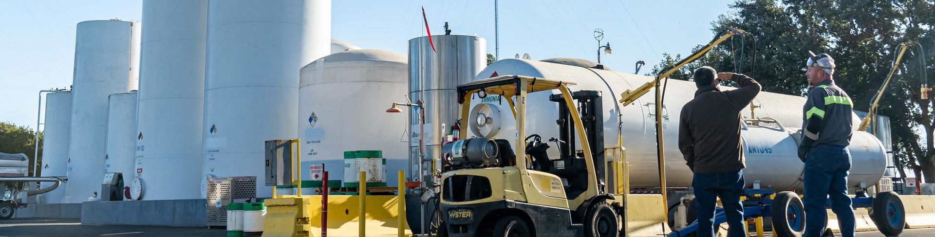 Men near industrial tanks with a forklift. One man wears work clothes.