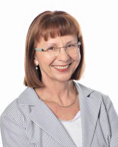 Woman with short auburn hair and glasses smiling; wearing a striped blazer and white top.