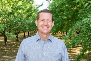Man in light blue shirt smiling in orchard. Trees in background.