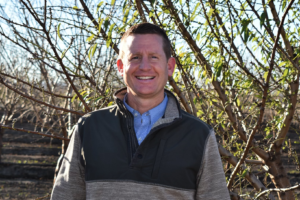 Man smiles outdoors in front of bare tree branches. He wears a blue shirt and green and gray sweater.