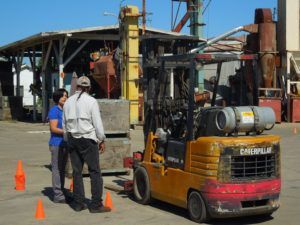 A yellow Caterpillar forklift carries a load; two people watch, with cones in a yard with industrial buildings.
