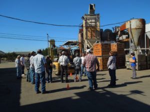 Group of people on a tour in front of industrial buildings, some are listening to someone speaking.