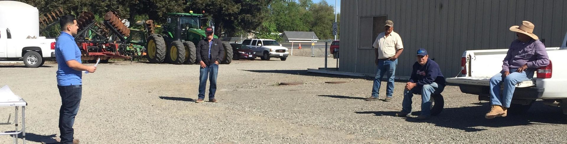 Group of people outdoors near a tractor and building, some are standing, others sitting in a truck bed, sunny day.
