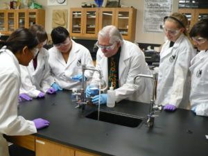 Students in lab coats watch instructor pour liquid into a beaker, sink in foreground.