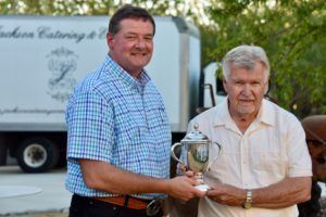 Man in blue shirt presents trophy to older man; catering truck in background.