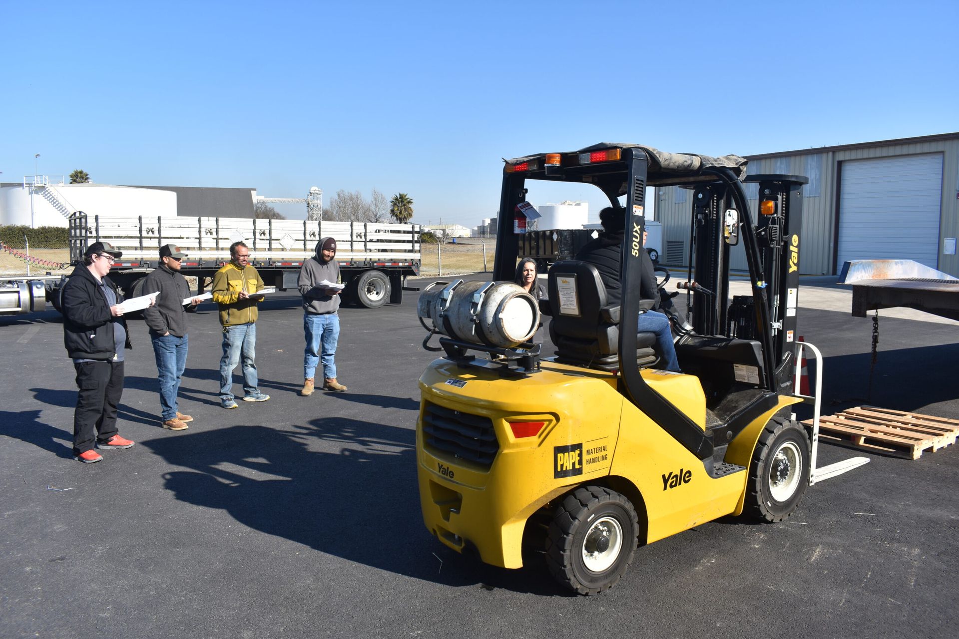Yellow forklift with operator; people observing. Outdoor setting.