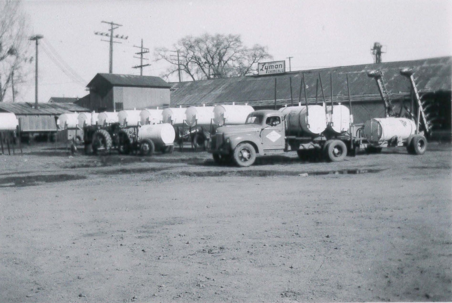 Vintage trucks with tanker trailers parked at a loading area; building in background.