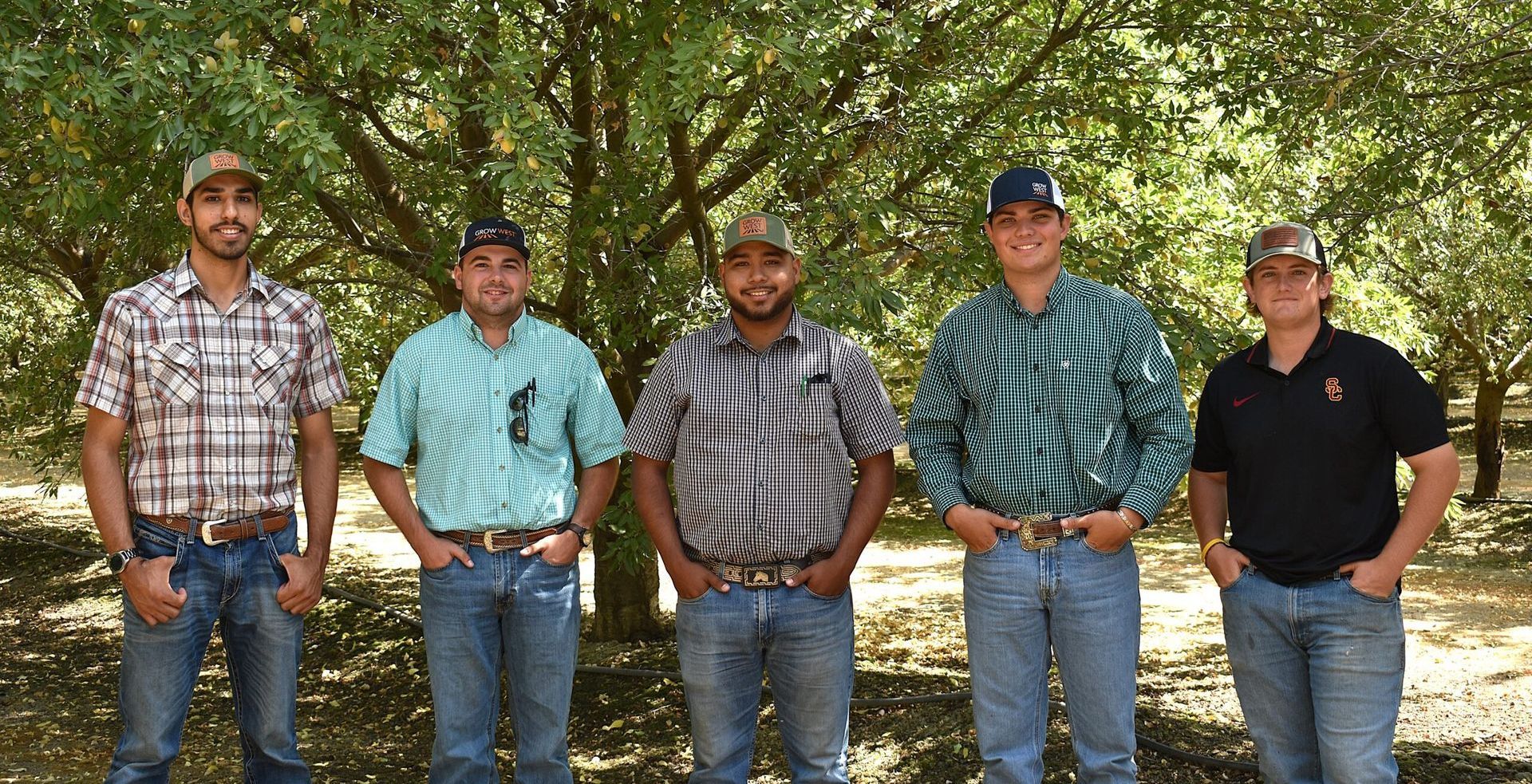 Five people standing in front of trees, wearing hats, shirts, and jeans.