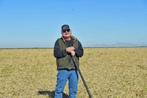 Man stands in a field, holding a dark stick. Blue jeans, green vest, and a cap. Clear blue sky.