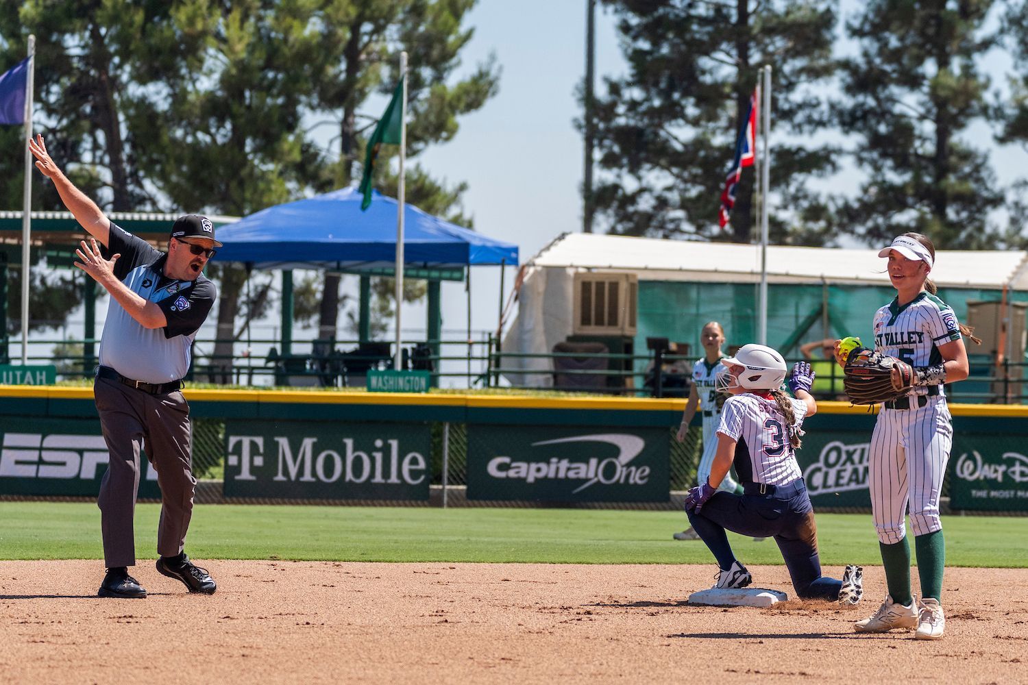 Softball game: Umpire signals safe as runner slides into base; fielder watches.