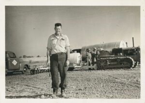 Man stands in front of machinery, possibly a tractor and truck, on an open field.