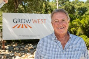 Man smiling in front of a GROW WEST sign; outdoors, trees in background.