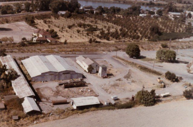 Aerial view of a nursery with several greenhouses, buildings, a road, and trees, with a lake in the background.