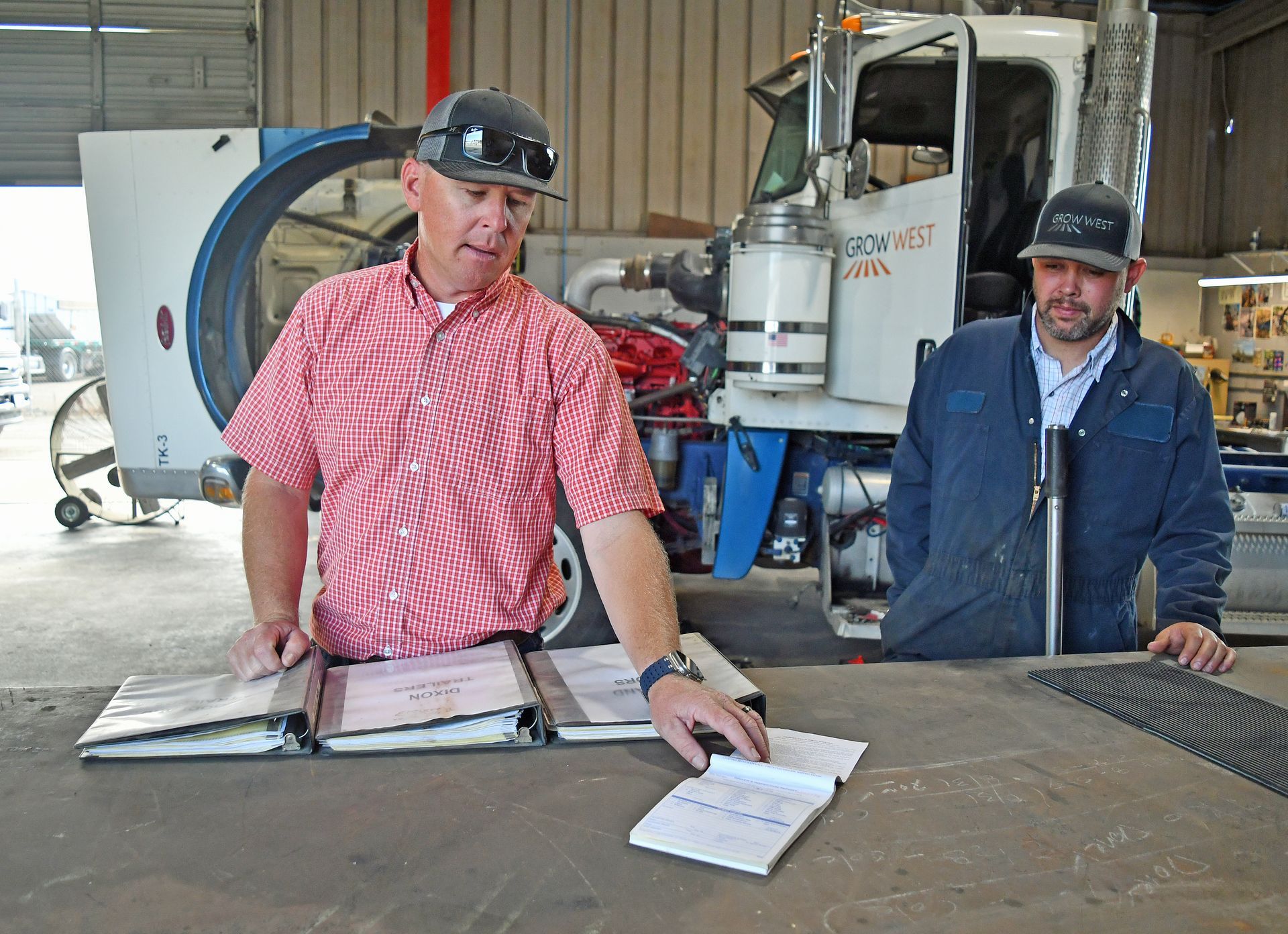 Two men reviewing documents near a large truck, one wearing a red shirt, the other a blue jacket, in an industrial setting.