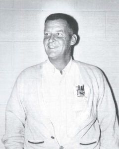 Man in a light-colored jacket smiles, looking to the side. A small logo is on the jacket.