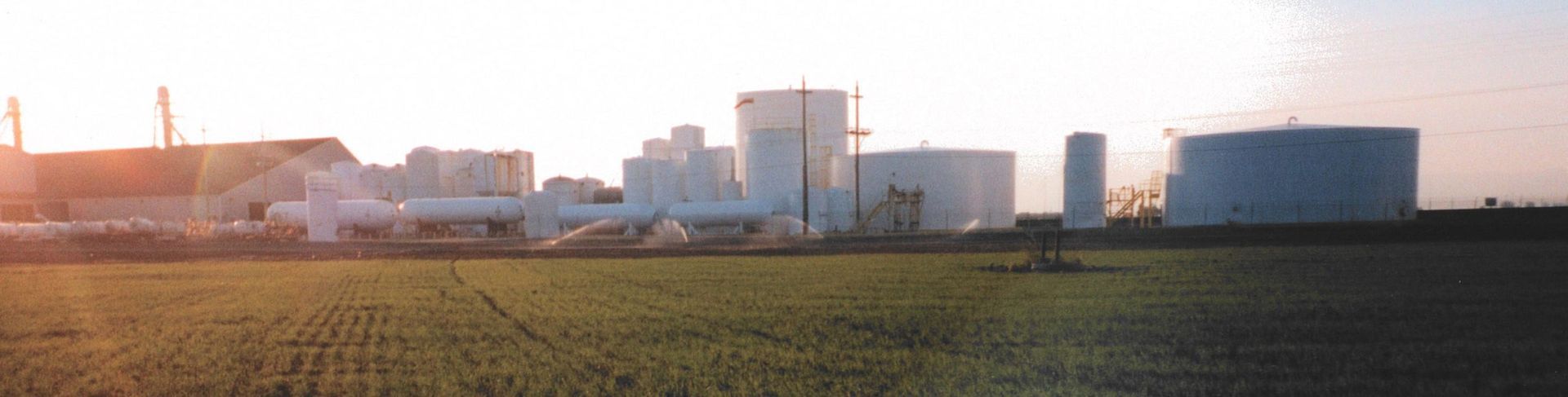 Industrial complex with silos and tanks in field under a bright sky.