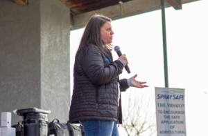 Woman speaking into microphone outdoors, gesturing, near sign about agricultural spraying.
