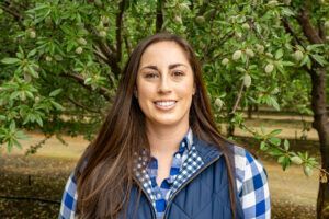 Woman smiling, wearing a blue plaid shirt and vest, standing in front of almond trees.