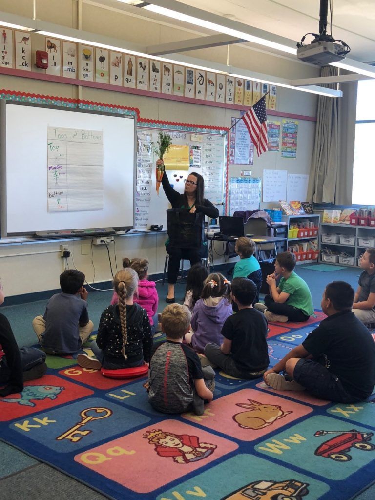 Teacher holding carrots in a classroom, students seated on the floor.