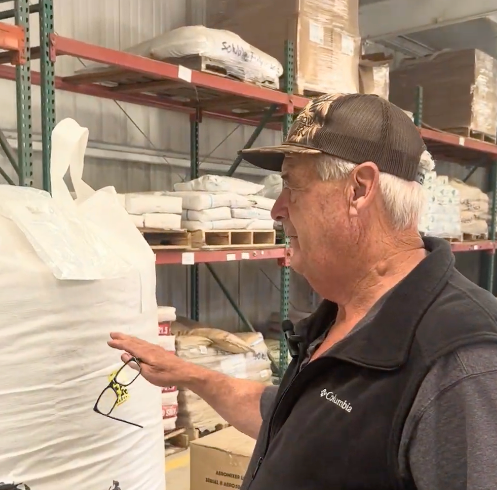 Man in a warehouse points at a large white bag, wearing a cap and vest. Shelves with goods in background.