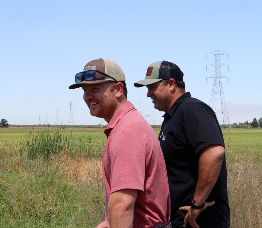 Two men in caps walking by tall green grass, under a blue sky, with power lines in the background.