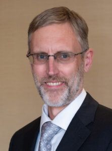 Man in glasses, suit, and tie smiling, soft background.