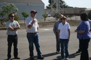 Group of people standing outside; man in hat speaks, others listen. Buildings and tower in background.