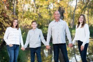 Family holding hands, posing in front of a tree-lined body of water. They wear casual clothing, smiles.