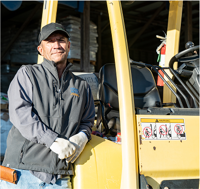 Man in work clothes leans on a yellow forklift. He wears a hat and gloves, smiling slightly. Warehouse setting.
