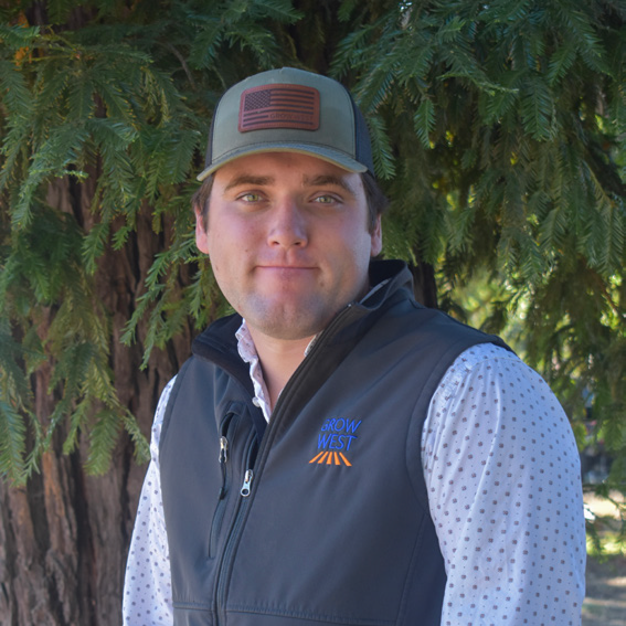 Man wearing a hat and vest smiles, standing outside near a tree.