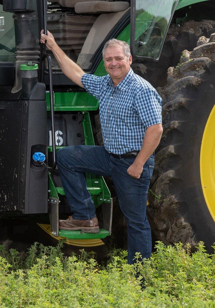 Man in blue shirt and jeans steps onto a John Deere tractor in a field.