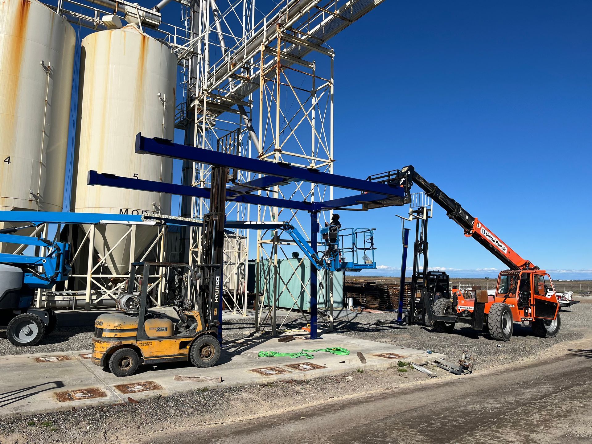 Construction site with forklift, crane, and workers assembling a blue metal structure near storage tanks.