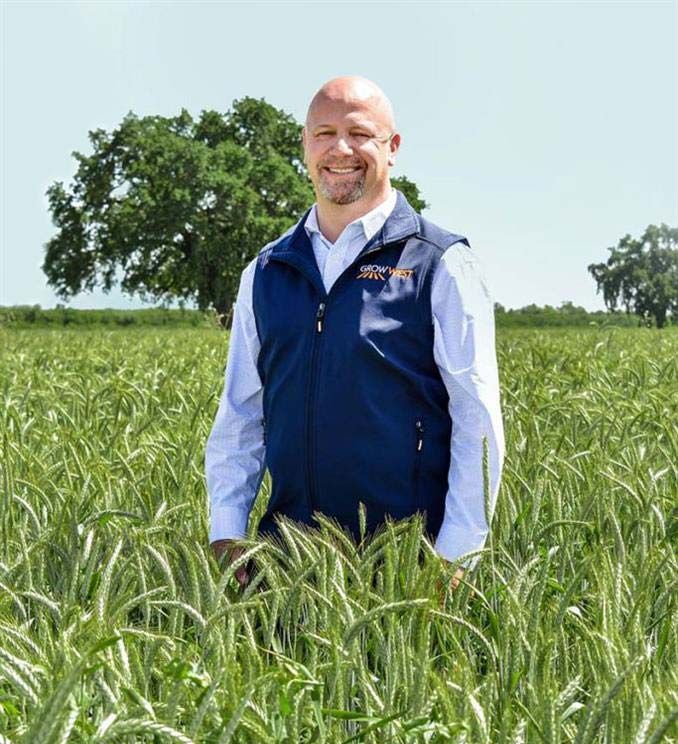 Man standing in a field of wheat, wearing a blue vest and white shirt, smiling.