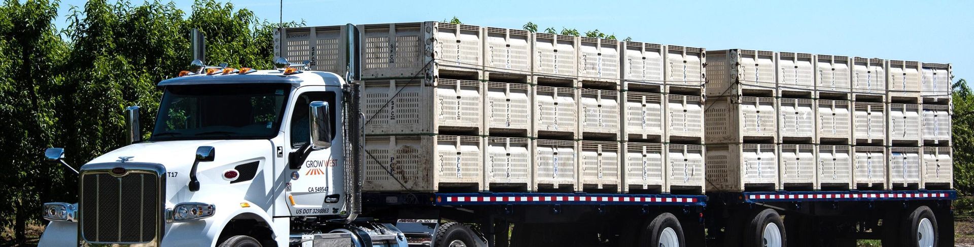 A white truck hauling stacked wooden crates on a trailer, trees in the background.