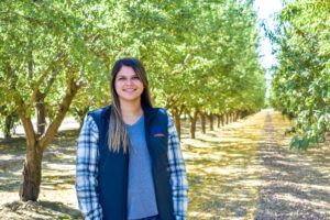 Woman in plaid shirt and vest smiles in an orchard with rows of trees.