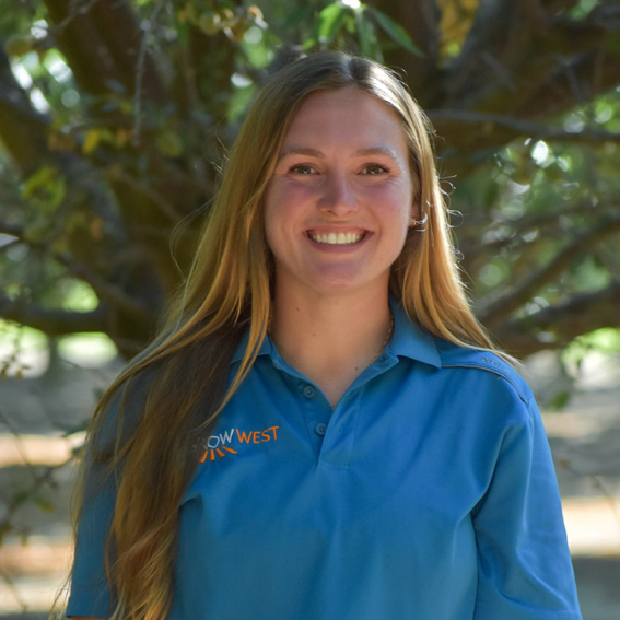 Woman with long blonde hair, smiling, wearing a blue polo shirt, tree background.