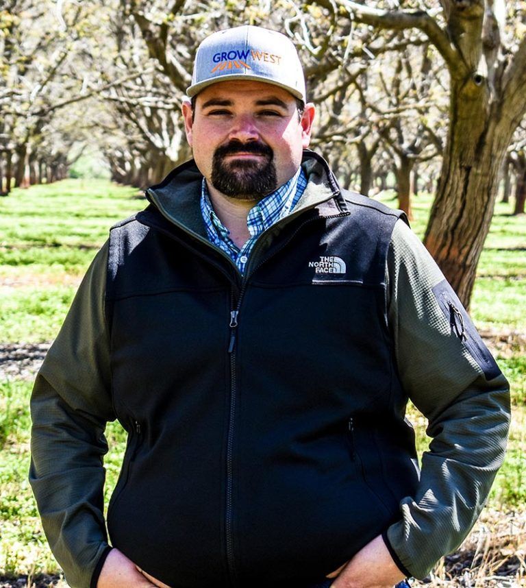 Man in a vest and baseball cap in an orchard. He has a beard and hands in his pockets.