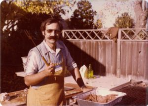 Man with mustache grilling, holding tools; outdoor setting with fence and foliage.