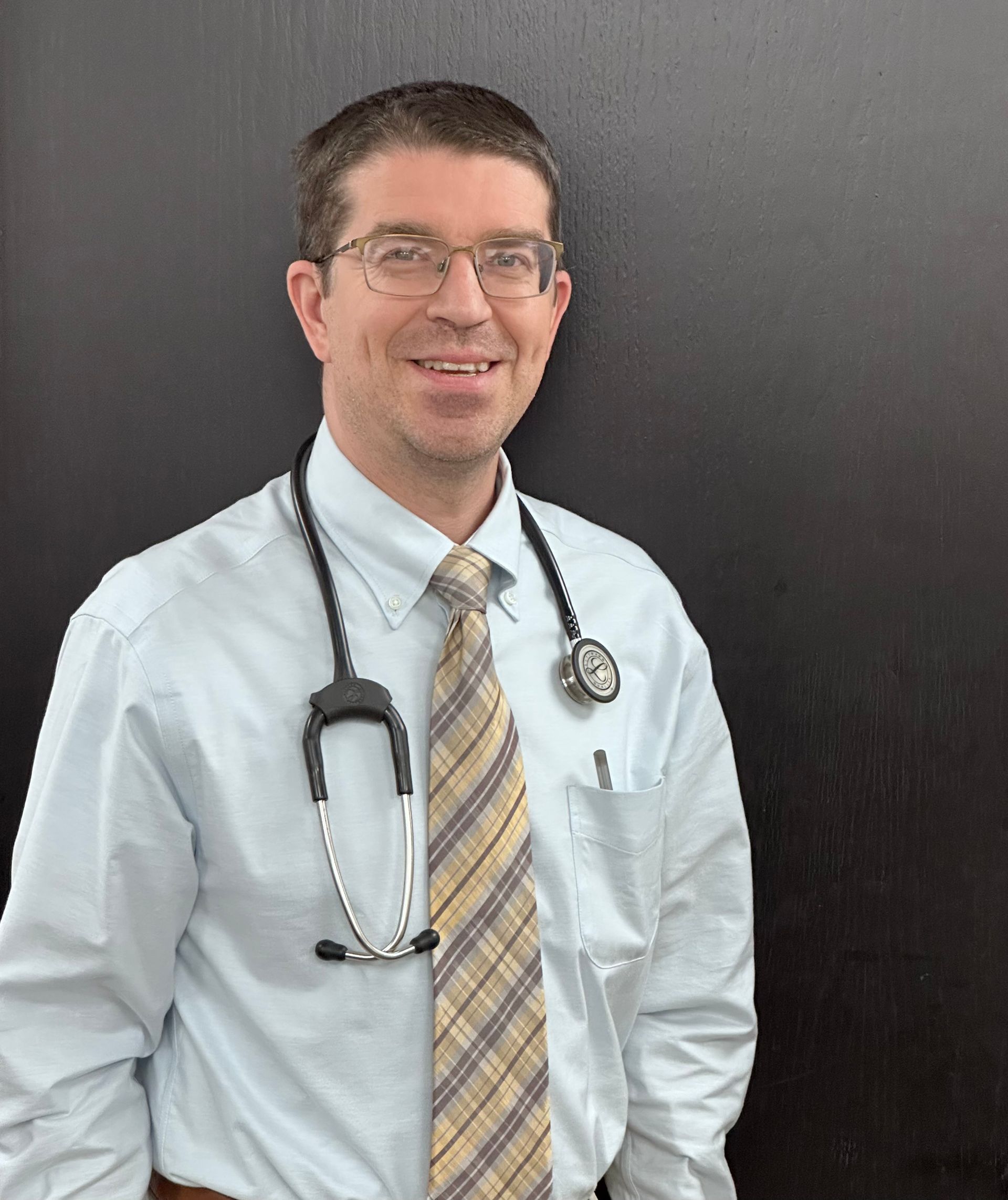 Doctor with stethoscope smiling, wearing glasses, checked shirt, and tie.
